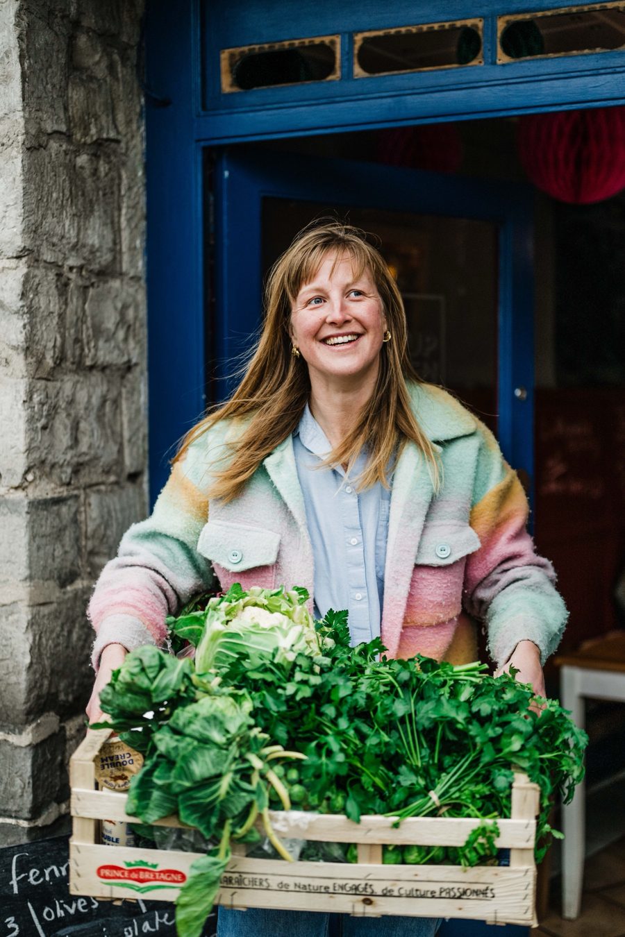 Annie Lobster, private chef in Barcelona, smiling in a doorway, carrying a crate of cauliflower, parsley and greens